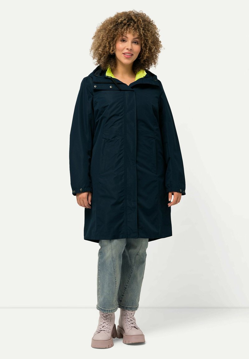 Cappotto impermeabile navy con cappuccio, foderato internamente di un vivido verde, tasche frontali e una vestibilità comoda. Abbinato a jeans azzurri e stivali rosa.