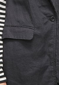 Veste en lin noire avec une poche poitrine, tissu texturé, et un bouton visible. Un tissu rayé d'une chemise dépasse en dessous.