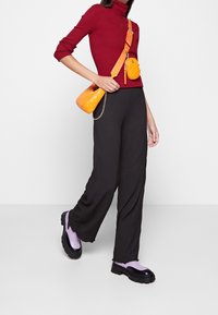 Col roulé rouge côtelé, pantalon large texturé noir, chaussures à plateforme noires avec accents lavande ; sac bandoulière matelassé jaune sur l'épaule.