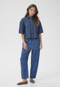 Denimjakke med korte ærmer og brystlommer, parret med løstsiddende denimbukser. Begge er i en medium blå vask med synlig syning.