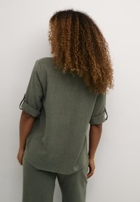 Donna con lunghi capelli ricci, che indossa una maglia verde oliva con maniche arrotolate e pantaloni abbinati, di fronte a una parete grigia chiara.