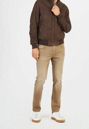 Homme portant une veste en cuir marron, un jean beige et des baskets blanches, debout devant un fond clair uni.