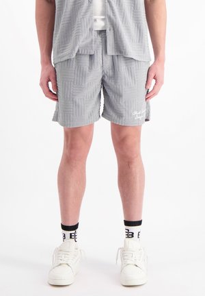 Grijs gemusterde shorts met een textuurdesign, voorzien van een wit geborduurd logo, gecombineerd met witte sneakers en zwarte gemusterde sokken.