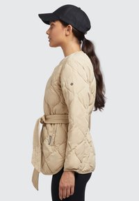 Vrouw in een beige gewatteerde jas met riem en zwarte pet, afgebeeld in profiel tegen een effen achtergrond.