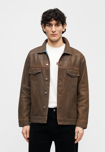 Καφέ δερμάτινο μπουφάν με γιακά, δύο τσέπες στο στήθος και κουμπώματα με κουμπιά. Φορεμένο πάνω από ένα λευκό πουκάμισο, με μπλε φόδρα ορατή στον γιακά.