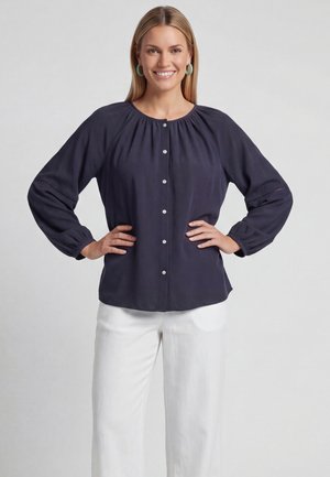 Vrouw glimlachend met handen in haar zij, gekleed in een marineblauw blouse met lange mouwen en witte broek tegen een effen achtergrond.