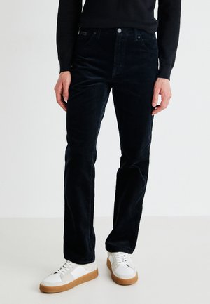 Pantalon en velours côtelé bleu marine avec une coupe droite, comportant cinq poches, une fermeture à bouton, porté avec des baskets blanches.