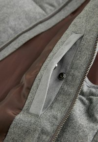 Wnętrze szarej kurtki z tkaniny z brązowym podszewką oraz kieszenią na zamek błyskawiczny. Do wyróżniających się detali należy guziczek nap i gładka tekstura.