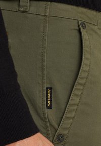 Pantalon cargo vert olive avec une texture lisse, mettant en avant une étiquette noire avec un texte jaune, et un bouton visible sur la taille.