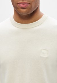 Mann trägt einen cremefarbigen Rundhals-Pullover mit kleiner quadratischer "BOSS"-Logo-Patch auf der linken Brust, neutraler Hintergrund.