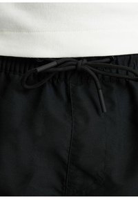 Pantaloni corti neri in tessuto testurizzato, con una vita elasticizzata e un cordino con termini in plastica. Orlo di una camicia bianca visibile sopra.