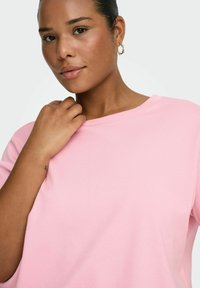 Jasnoróżowa bluzka z krótkim rękawem i okrągłym dekoltem, o miękkiej fakturze. Osoba trzyma się za szyję, prezentując proste kolczyki w kształcie koła.