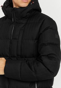 Schwarze Steppjacke mit Kapuze, ausgestattet mit einem Fischgrätenmuster, Reißverschluss und einer Brusttasche. Mit roten Ziehlaschen akzentuiert.
