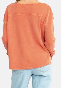 Pull à manches longues, coupe décontractée, en tissu texturé orange, avec un large décolleté et des épaules tombantes. Associé à un jean bleu clair.