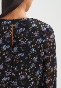Donna che indossa una blusa nera con stampa floreale viola e blu, apertura a chiave con bottoni sulla parte superiore della schiena e maniche lunghe trasparenti.