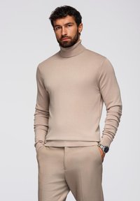 Maglione beige a collo alto con maniche lunghe, design slim fit, texture morbida, abbinato a pantaloni coordinati, con un orologio al polso.