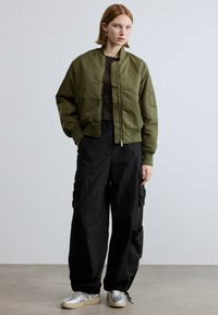 Χακί bomber μπουφάν με ριμπ μανσέτες και γιακά, σε συνδυασμό με φαρδιά μαύρα cargo παντελόνια. Τα παπούτσια είναι ασημένια με ανοιχτή σόλα.