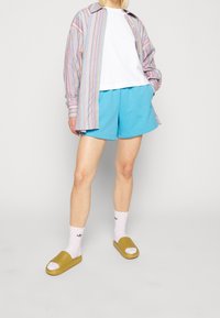 Ränder långärmad skjorta, vit t-shirt, blå shorts, vita strumpor, gula tofflor. En avslappnad outfit med olika färger och texturer.