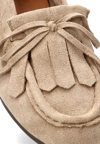 Acercamiento de un mocasín de ante beige con flecos y detalles de lazada en la parte superior, destacando el material texturizado y la punta redondeada.