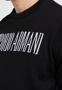 Svart tröja med hög krage, stickad textur och framträdande vitt "EMPORIO ARMANI"-logotyp över bröstet.