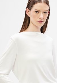 Fehér, hosszú ujjú felső magas nyakkal és puha textúrával. Egyszerű kialakítása látható minták vagy díszítések nélkül.