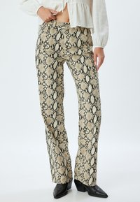 Pantalons à motif serpent beige et noir avec une coupe évasée, fabriqués en tissu texturé, associés à des bottes à talons aiguilles noires.
