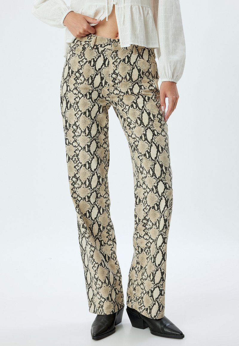 Pantalons à motif serpent beige et noir avec une coupe évasée, fabriqués en tissu texturé, associés à des bottes à talons aiguilles noires.