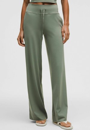 Kobieta w oliwkowozielonych spodniach o szerokich nogawkach z sznurkiem w talii i beżowych sandałach, stojąca na jednolitym tle.