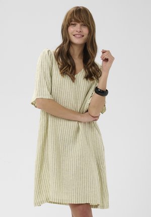 Donna con lunghi capelli castani che indossa un abito largo a righe verde chiaro e un bracciale nero, sorridente con le braccia incrociate su sfondo neutro.