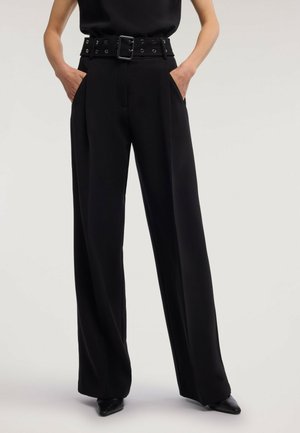Pantalon noir à jambes larges avec des plis et une large ceinture, porté les mains dans les poches et chaussures noires pointues visibles.