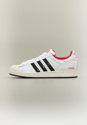 Λευκό αθλητικό παπούτσι Adidas με μαύρες ρίγες, κόκκινο εσωτερικό, λογότυπο Coca-Cola στη φτέρνα και λαστιχένια μύτη, εμφανιζόμενο από την εξωτερική πλευρά.