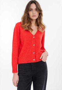 Červený pletený cardigan s výstřihem do V, sedmi kovovými knoflíky a žebrovanými manžetami a lemem. Nosený přes tmavé džíny, s uvolněným střihem.