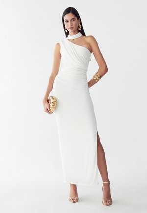 Frau in einem weißen Ein-Schulter-Kleid mit einem goldenen Manschettenarmband, einer goldenen, muschelförmigen Clutch und goldenen High-Heeled Sandalen steht vor weißem Hintergrund.