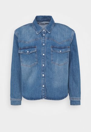 Chemise en denim bleu à boutons, manches longues, deux poches à rabat avec pressions à l'avant, et col pointu, présentée sur un fond blanc.