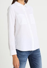 Kvinde iført en hvid skjorte med lange ærmer og vertikale striber samt blå jeans, stående mod en ensfarvet baggrund.