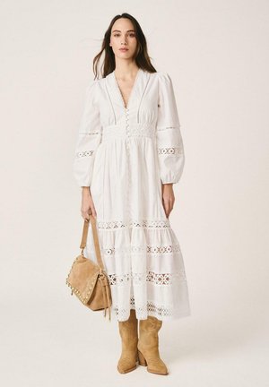 Mujer con vestido midi de encaje blanco de manga larga, sosteniendo un bolso de mano de ante marrón claro y luciendo botas de tacón de ante del mismo color, posando frente a un fondo neutro.