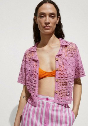 Rosa gehäkelte Kurzarmkarte mit floralen Mustern und Holzknöpfen, über einem orangefarbenen Crop-Top getragen, kombiniert mit gestreiften Hosen.