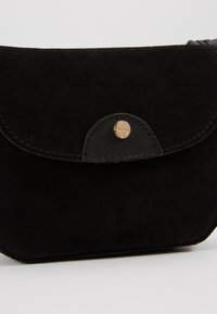 Pochette en daim noir avec un rabat arrondi sécurisé par un petit bouton rond en or sur une surface blanche lisse.