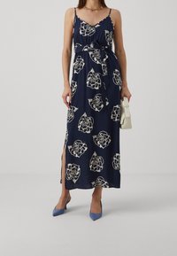 Kvinna i en marinblå blommig ärmlös maxiklänning med midjeband, blå spetsiga klackar och håller en liten vit handväska.