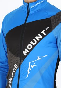 Blauwe fietsjack met een zwart geometrisch patroon, ritssluiting en wit logo. Heeft een gestructureerde stof en ademende ontwerpelementen.