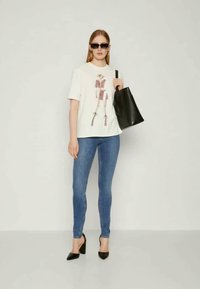 Λευκό μπλουζάκι με γραφικό τύπο μιας φιγούρας μόδας, συνδυασμένο με μπλε στενά τζιν, μαύρες ψηλές γόβες και μία μαύρη τσάντα ώμου.