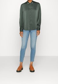 Ciemnozielona satynowa koszula z klasycznym kołnierzykiem, zapinana na guziki z przodu, z długimi rękawami; w połączeniu z jasnoniebieskimi obcisłymi dżinsami i brązowymi butami wsuwanymi.