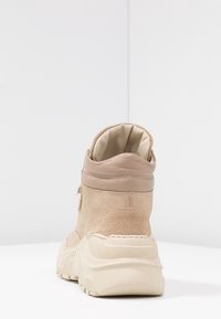 Zapatilla alta de ante beige con cuello acolchado y suela de goma gruesa. La vista trasera muestra un acento de cuero liso cerca de la parte superior.