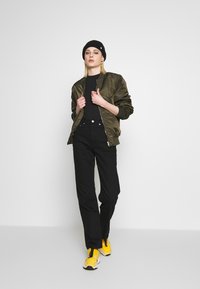 Casaco bomber verde-oliva, top preto ajustado, calças pretas de cintura alta, ténis amarelos, gorro preto, de pé contra um fundo branco.