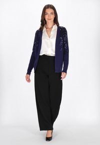 Navy Strickjacke mit Paillettenakzenten, getragen über einer weißen Bluse, kombiniert mit schwarzen weit geschnittenen Hosen und schwarzen High Heels. Insgesamt glatte Textur.