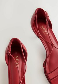 Zapatos planos rojos hechos de cuero suave, con una tira delgada con hebilla y un diseño de punta redondeada. Logo "NEXT" grabado en la plantilla.