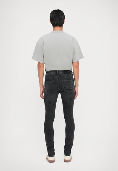 Camicia grigia a maniche corte abbinata a jeans skinny neri con dettagli strappati, due tasche posteriori e una silhouette aderente.
