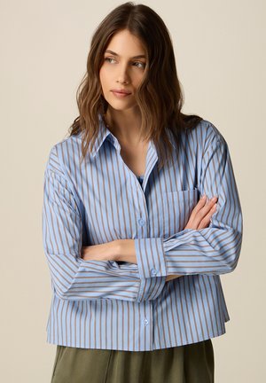 Giovane donna con capelli castano ondulati che indossa una camicia a righe blu e marrone con bottoni, braccia incrociate, che guarda di lato su uno sfondo beige.