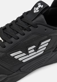 Chaussure de sport noire avec une tige en synthétique texturé, des accents de logo argentés et des lacets plats. Motif distinctif sur le panneau latéral et semelle robuste.
