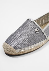 Chaussure slip-on argentée pailletée avec semelle beige tissée et petit logo métallique rond sur le côté supérieur, présentée sur fond blanc.
